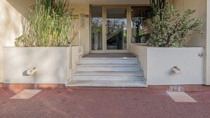 Een gebouwtoegang met een trap die leidt naar een glazen deur. Aan weerszijden van de ingang staan plantenbakken met verschillende planten.