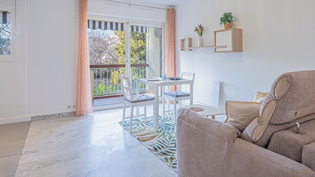 Een moderne woonkamer met een bank, een eettafel met stoelen, een balkon met uitzicht op bomen, en een hanglamp.