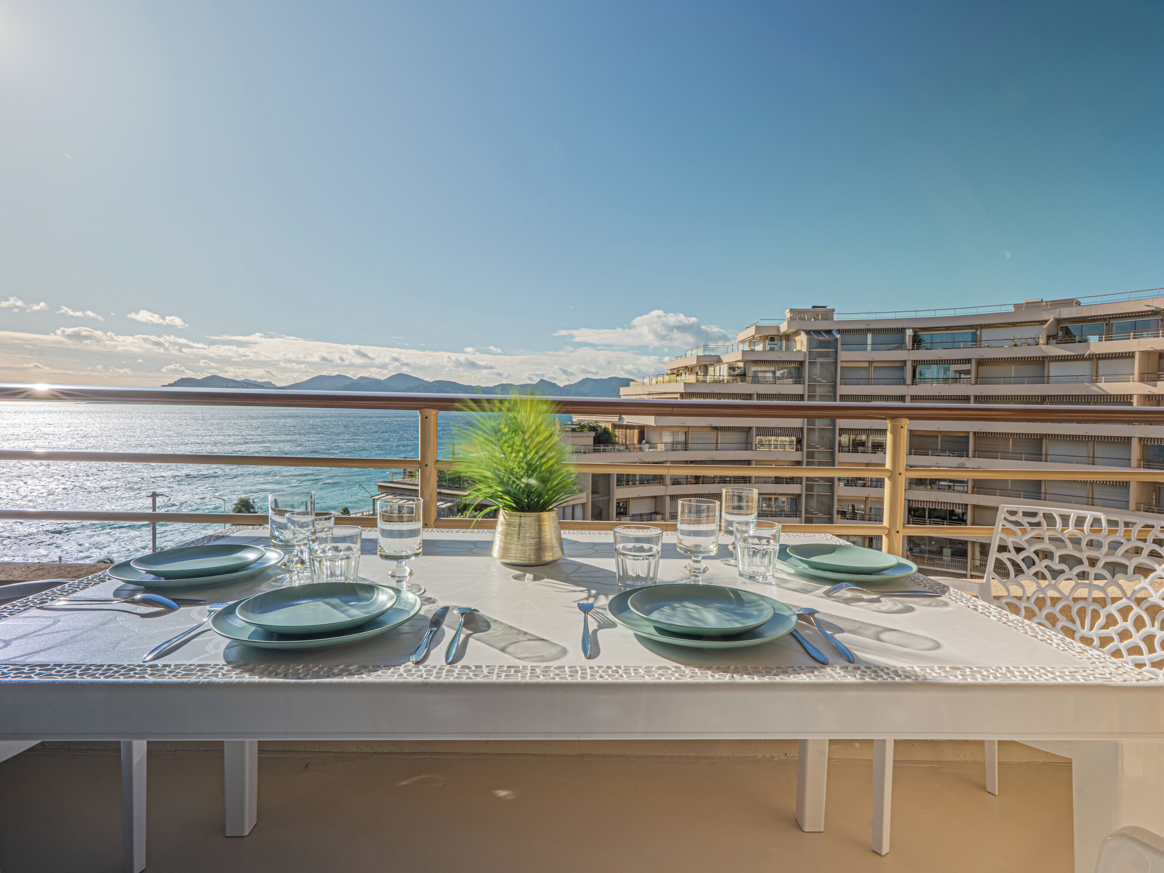 Ein Esstisch ist gedeckt mit Tellern, Gläsern und Besteck auf einem Balkon mit Blick aufs Meer und auf Gebäude.