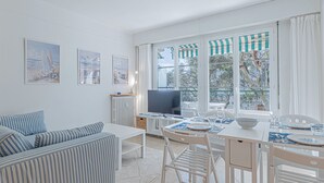 Uma sala iluminada com um sofá branco, uma mesa de jantar com cadeiras, uma TV e obras de arte nas paredes.