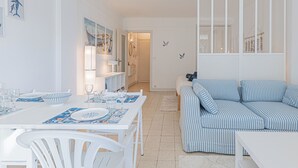 Um quarto claro e branco com uma mesa de jantar posta para quatro pessoas, um sofá listrado e vista para o mar através de uma janela.