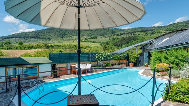 A swimming pool with a wooden chair and an umbrella, surrounded by stone tiles and greenery.