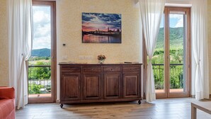 A room with a wooden cabinet, a painting of a landscape, and a view of greenery through large windows.