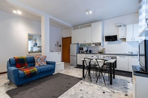 Eine moderne Küche mit weißen Schränken, einem Esstisch mit Stühlen und einem blauen Sofa mit bunter Decke.