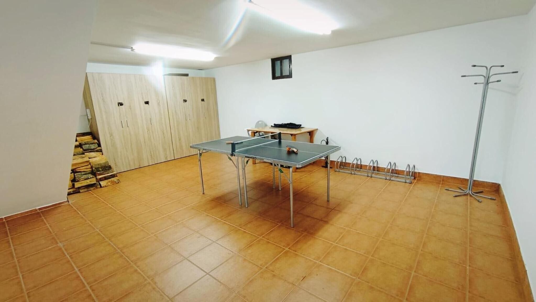 Una habitación con mesa de ping pong, perchero, pequeña mesa con sillas y una escalera.