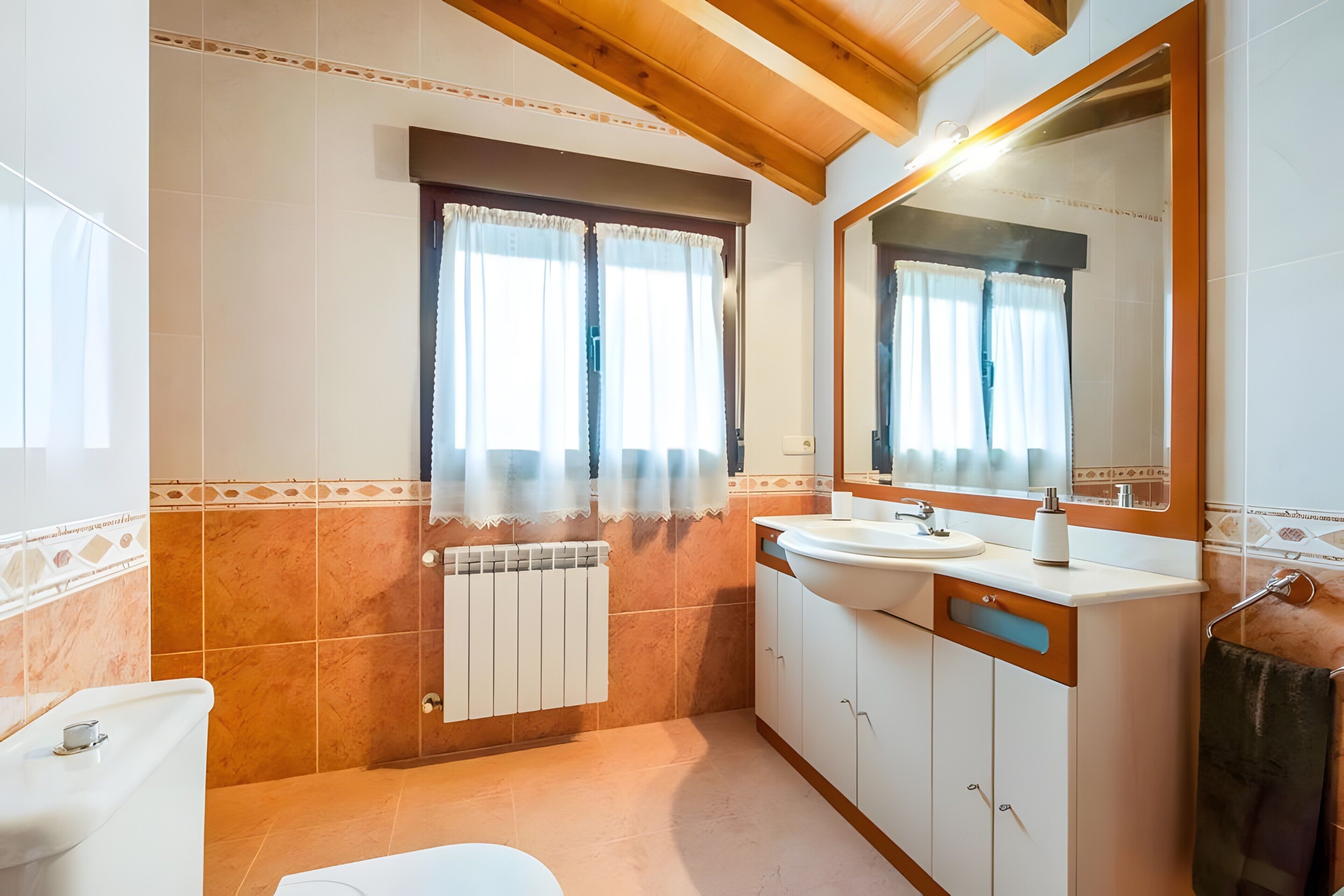 Une salle de bain avec un lavabo blanc, un miroir et des toilettes, des poutres en bois au plafond et une fenêtre avec des rideaux.