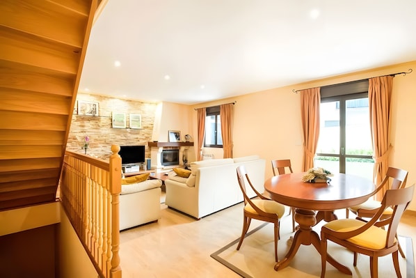 A modern living room with a dining area, wooden staircase, and a kitchen in the background.