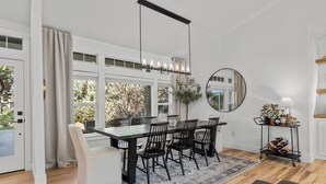 Un comedor con una mesa grande, sillas y un candelabro. Hay un espejo en la pared y una planta sobre un carrito.