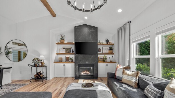 Una sala de estar moderna con chimenea, sofás grises, piso de madera y un ventanal con vistas a la vegetación.