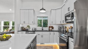 Una cocina moderna con armarios blancos, una isla grande, electrodomésticos de acero inoxidable y vistas al exterior a través de una ventana.