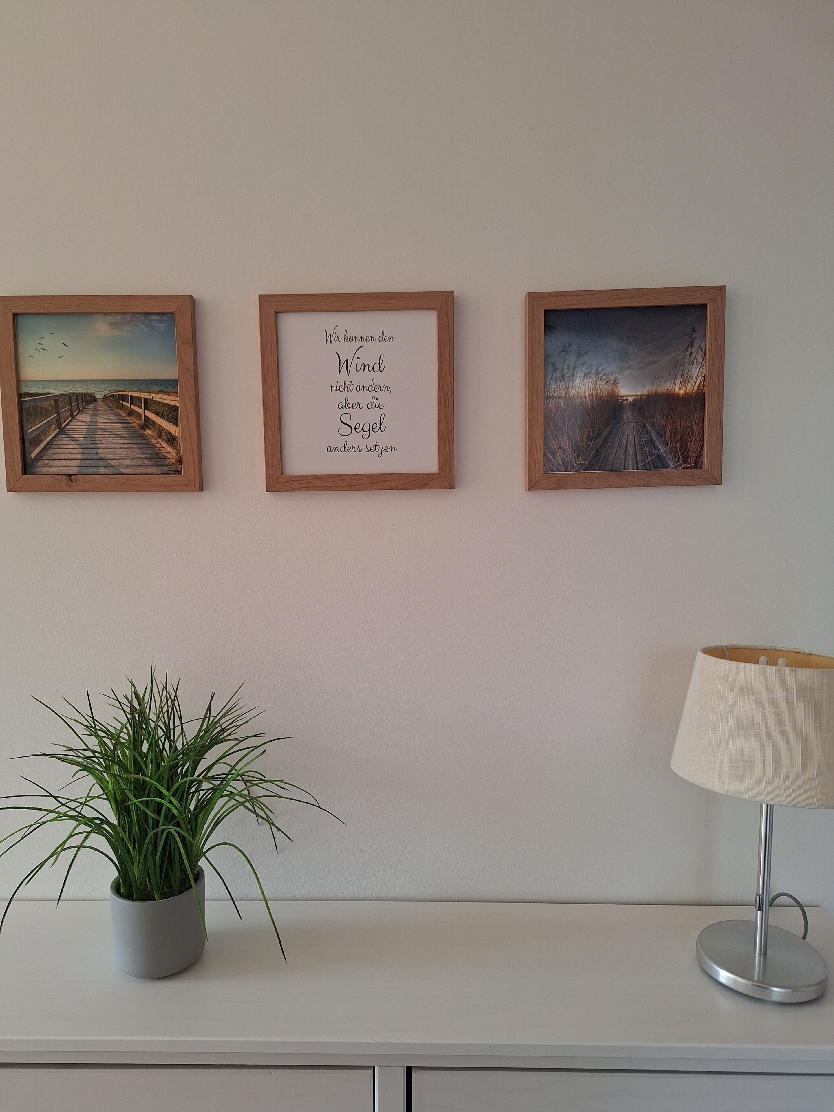 Drei gerahmte Bilder an einer Wand, ein Blumentopf und eine Lampe auf einem Tisch.