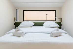 A double bed with white linens and towels, a green headboard, a window with blinds, and a wall-mounted light fixture. - Spacious Paris Duplex for 4 Near Bastille (Paris)
