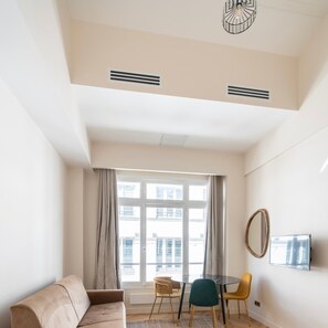 A modern living room with a sofa, dining table, chairs, a TV, and a large window with curtains. - Spacious Paris Duplex for 4 Near Bastille (Paris)