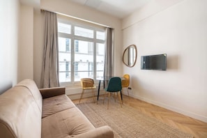 A living room with a sofa, a dining table with chairs, a TV mounted on the wall, and a window with curtains. - Spacious Paris Duplex for 4 Near Bastille (Paris)