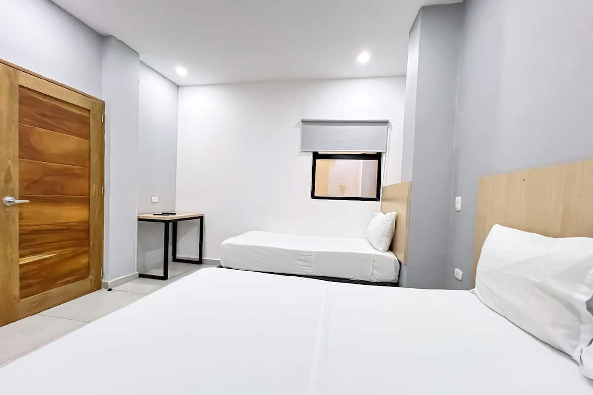A bedroom with a wooden door, a small table, a bed with white bedding, and a window with a view.