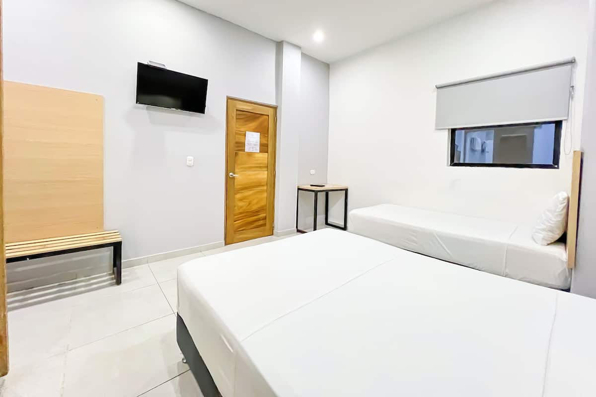 A modern hotel room with a large bed, a wooden bench, a small table, a TV mounted on the wall, and a window with a blind.