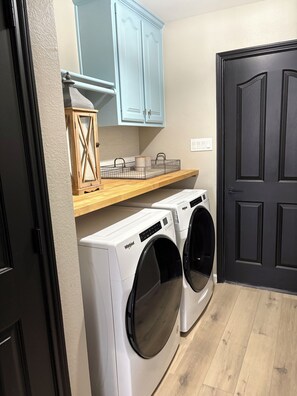 Un lavadero con dos lavadoras Whirlpool de carga frontal, un armario de madera con una cesta, y una puerta negra.