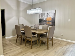 Un comedor con una mesa de madera y seis sillas tapizadas, una lámpara de araña y un cuadro con un paisaje urbano en la pared.