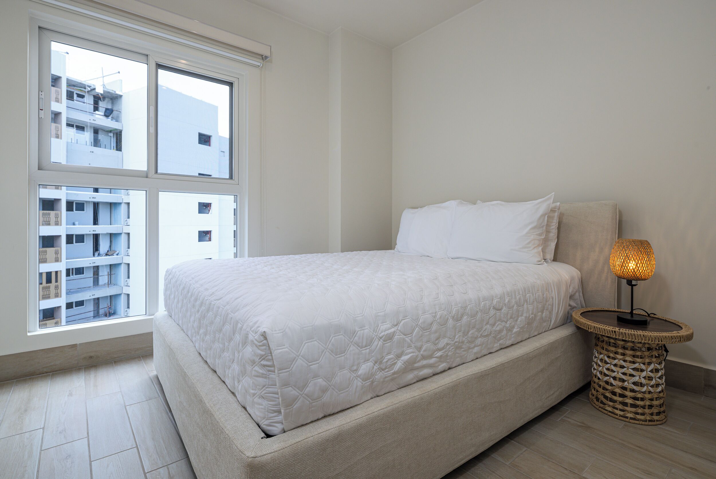 Une chambre à coucher avec un lit, une lampe de chevet et une fenêtre donnant sur des immeubles.