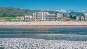 Un hotel frente a la playa con varias construcciones, una playa de arena y un paisaje montañoso de fondo.