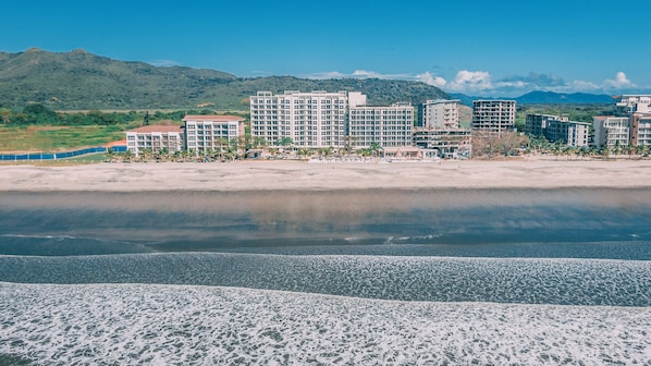 Ein Hotel direkt am Strand mit mehreren Gebäuden, einem Sandstrand und einer Bergkulisse.