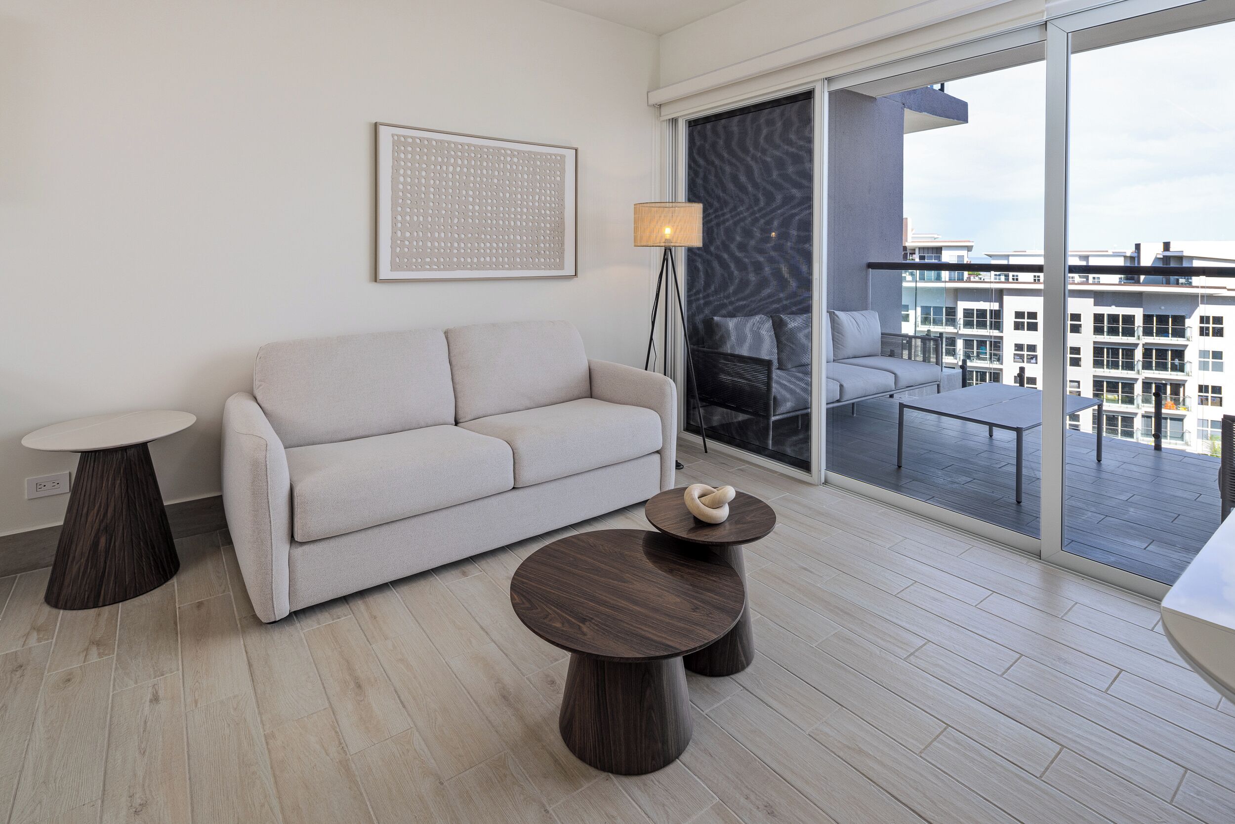 A modern living room with a sofa, two round tables, a floor lamp, and a large window with a view of buildings.