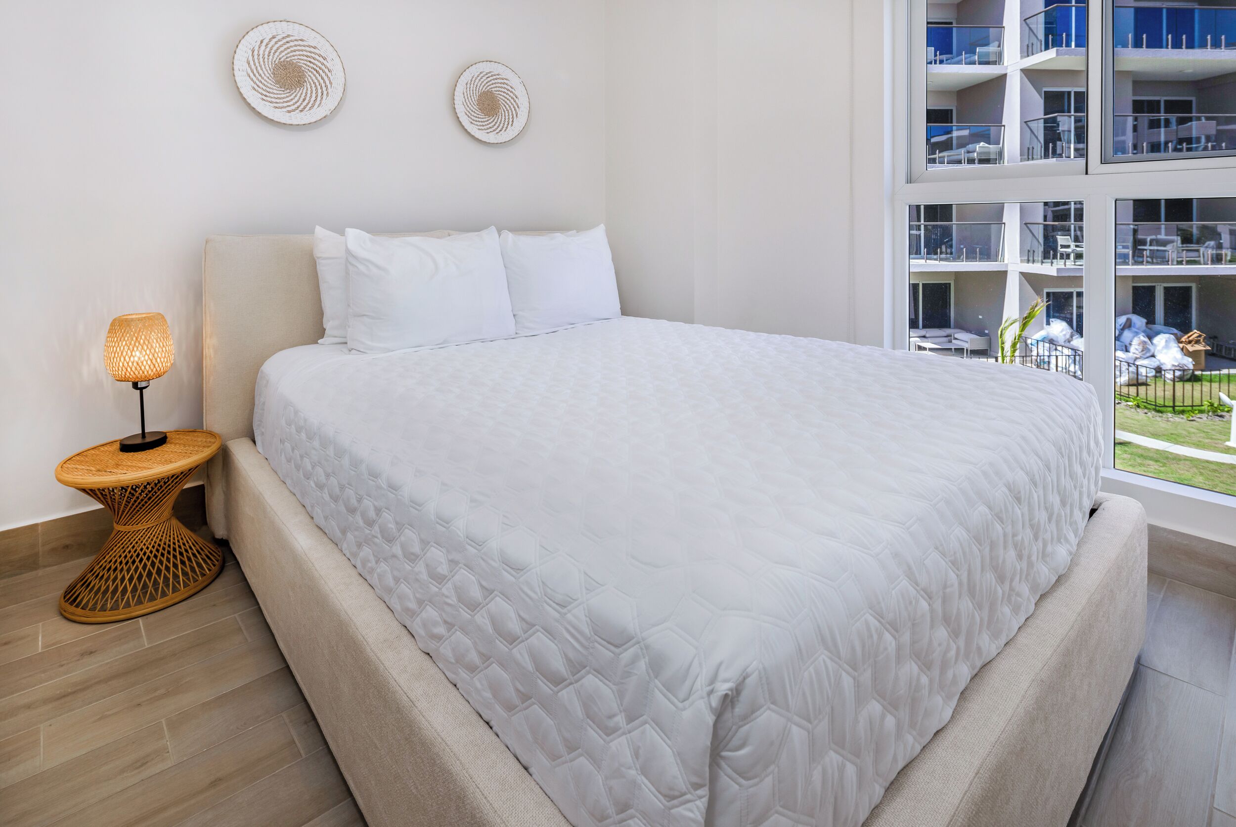 A modern bedroom with a large bed, a bedside lamp, and a view of a building with balconies.