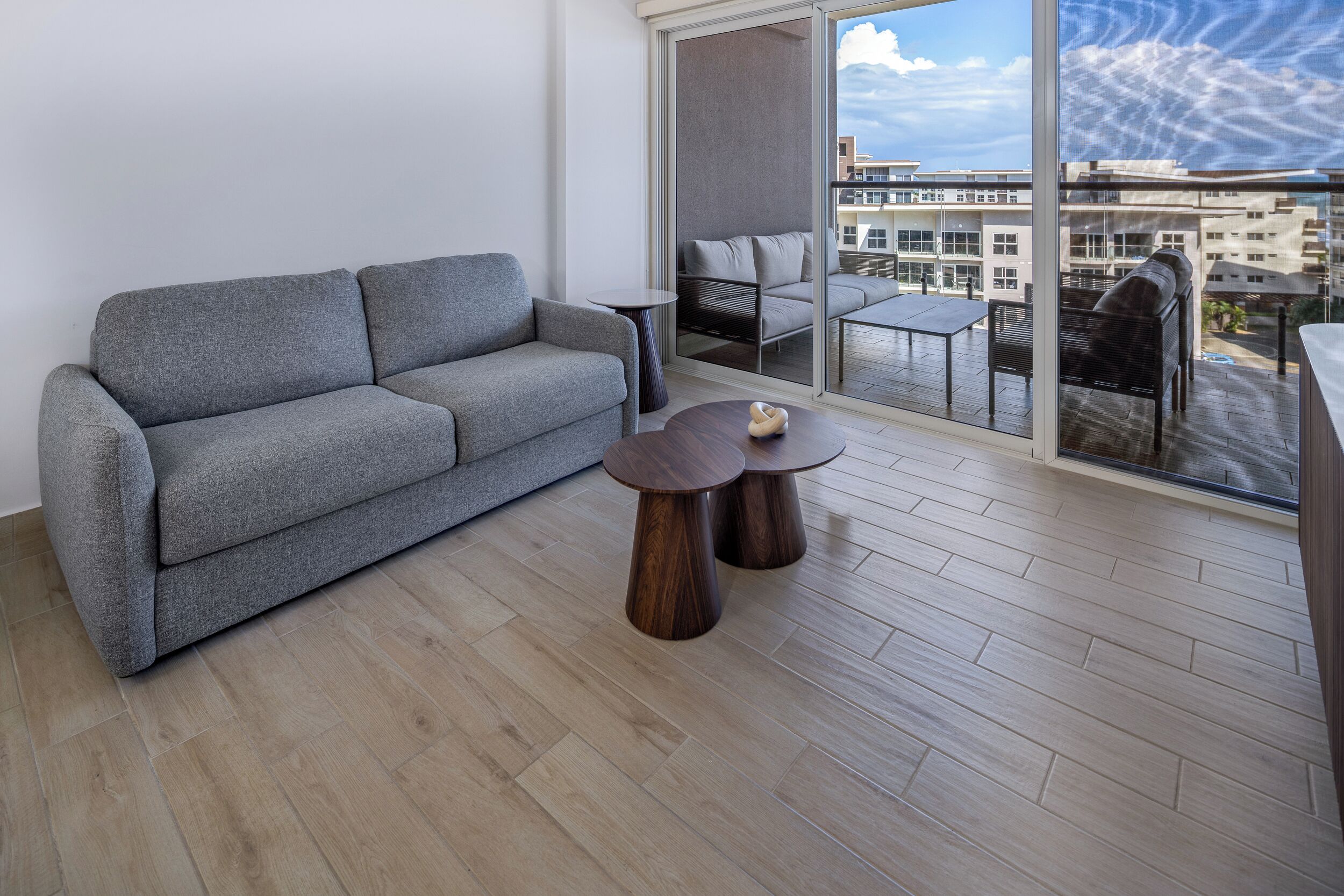 Un salon moderne avec un canapé gris, deux petites tables d’appoint en bois et une grande porte-fenêtre coulissante donnant sur un balcon extérieur avec vue sur la ville.