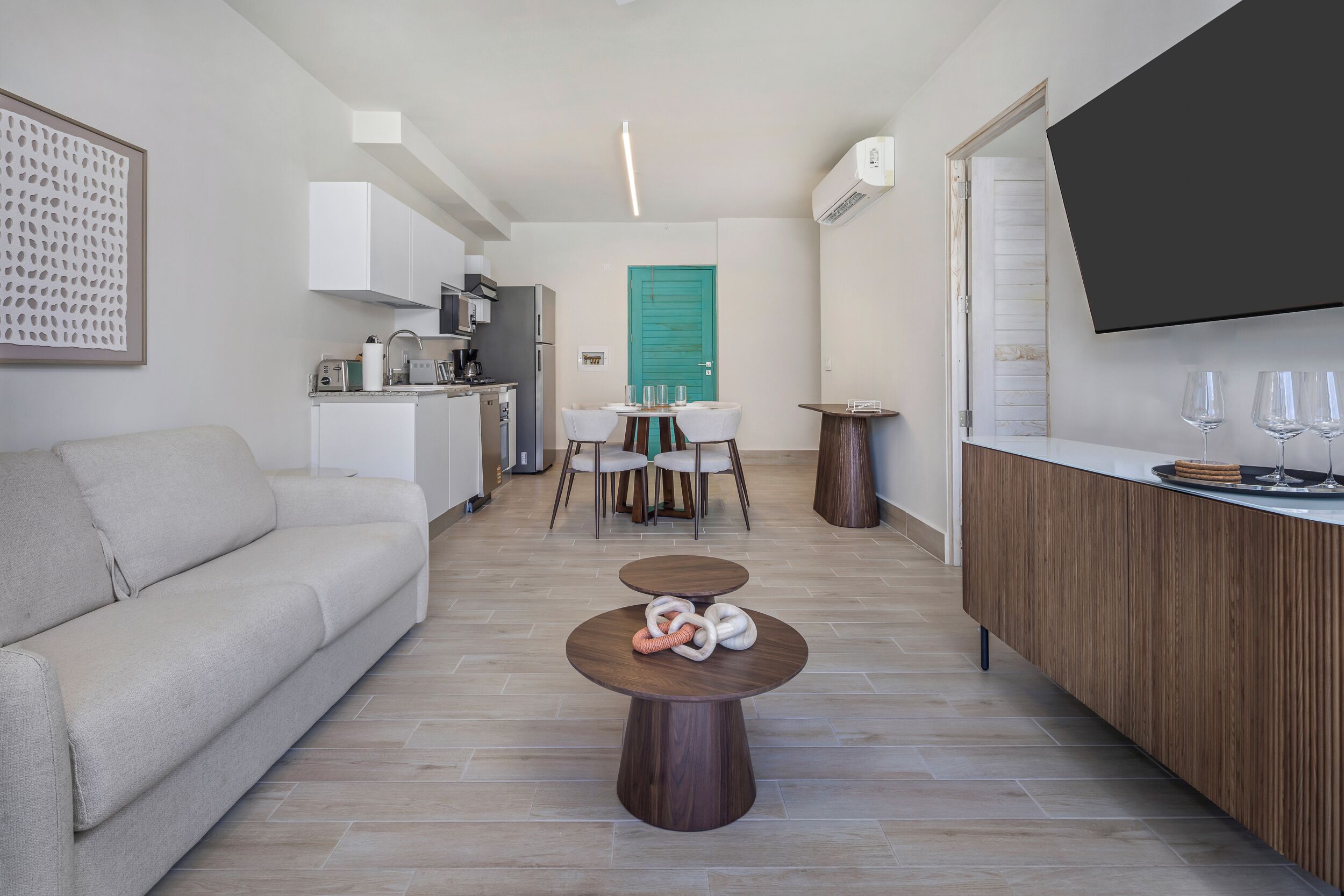 Un salon moderne comprenant un canapé, une table basse agrémentée d’objets décoratifs, un coin repas avec une table et des chaises, une cuisine équipée et un téléviseur fixé au mur.
