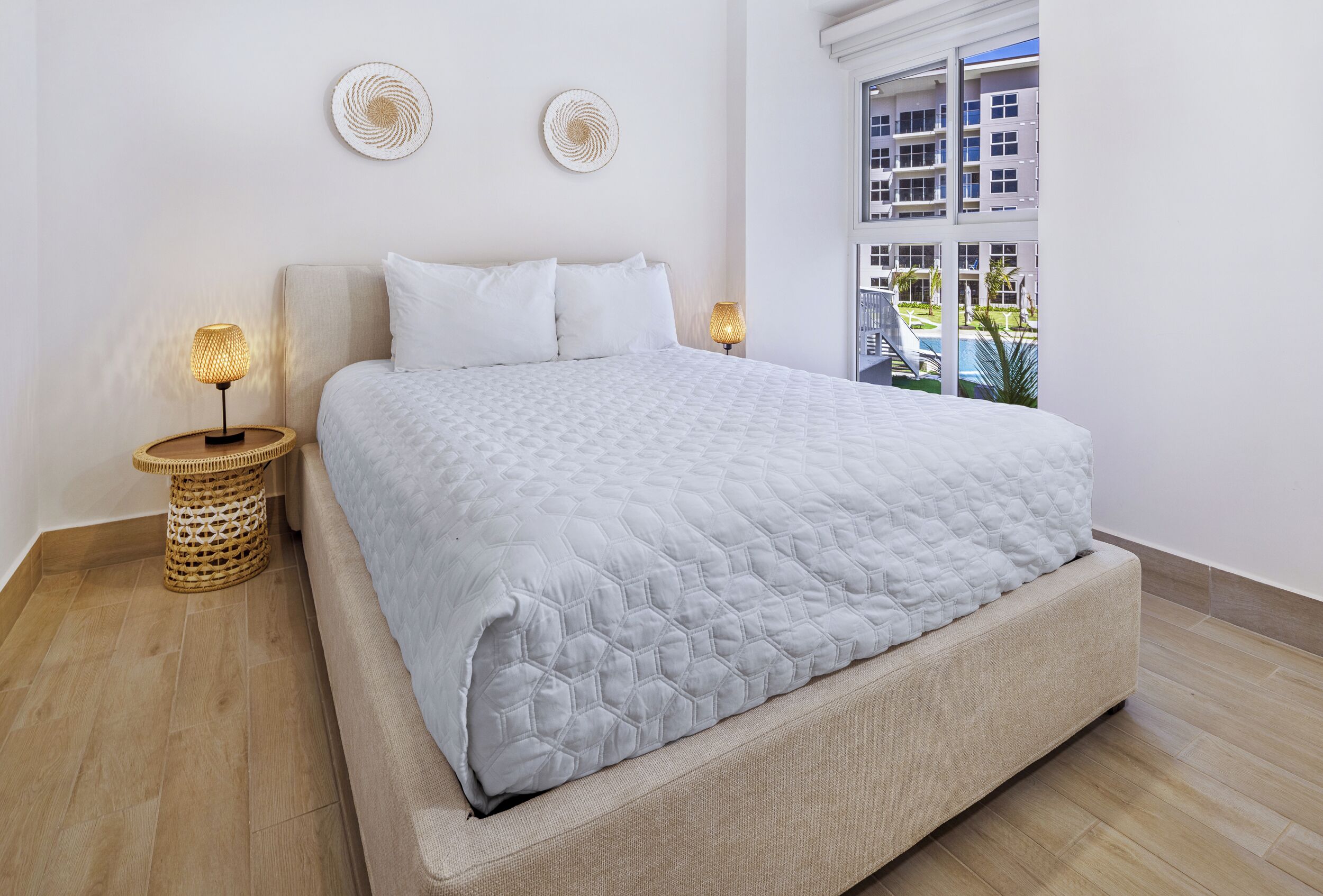 Una cama bien tendida con un edredón blanco acolchado, dos lámparas de noche y una mesita de noche tejida.