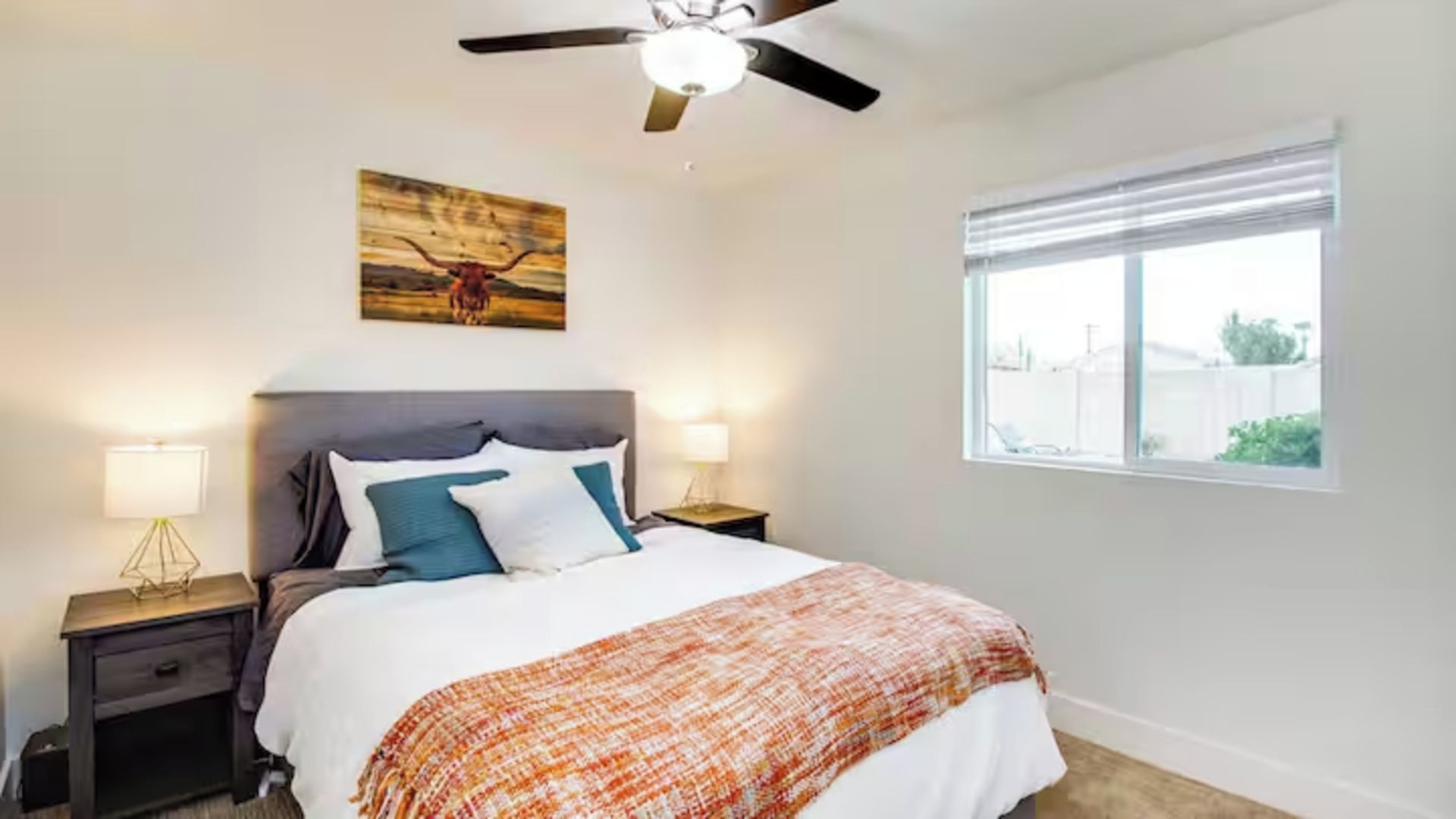 A bedroom with a bed, two bedside tables, a ceiling fan, a window with blinds, and a painting on the wall.
