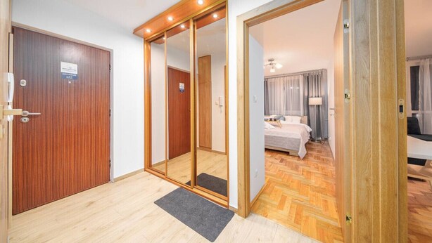 Una habitación de hotel con piso de madera, una cama con cubrecama blanco, una mesita de noche, una lámpara y vistas a un paisaje urbano a través de una ventana con cortinas.