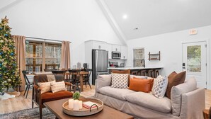 Uma sala de estar moderna com sofá, mesa de centro e árvore de Natal. Há uma área de cozinha com eletrodomésticos e uma mesa de jantar.