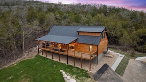 Uma cabana de toras de dois andares com varanda, rodeada por uma paisagem arborizada.