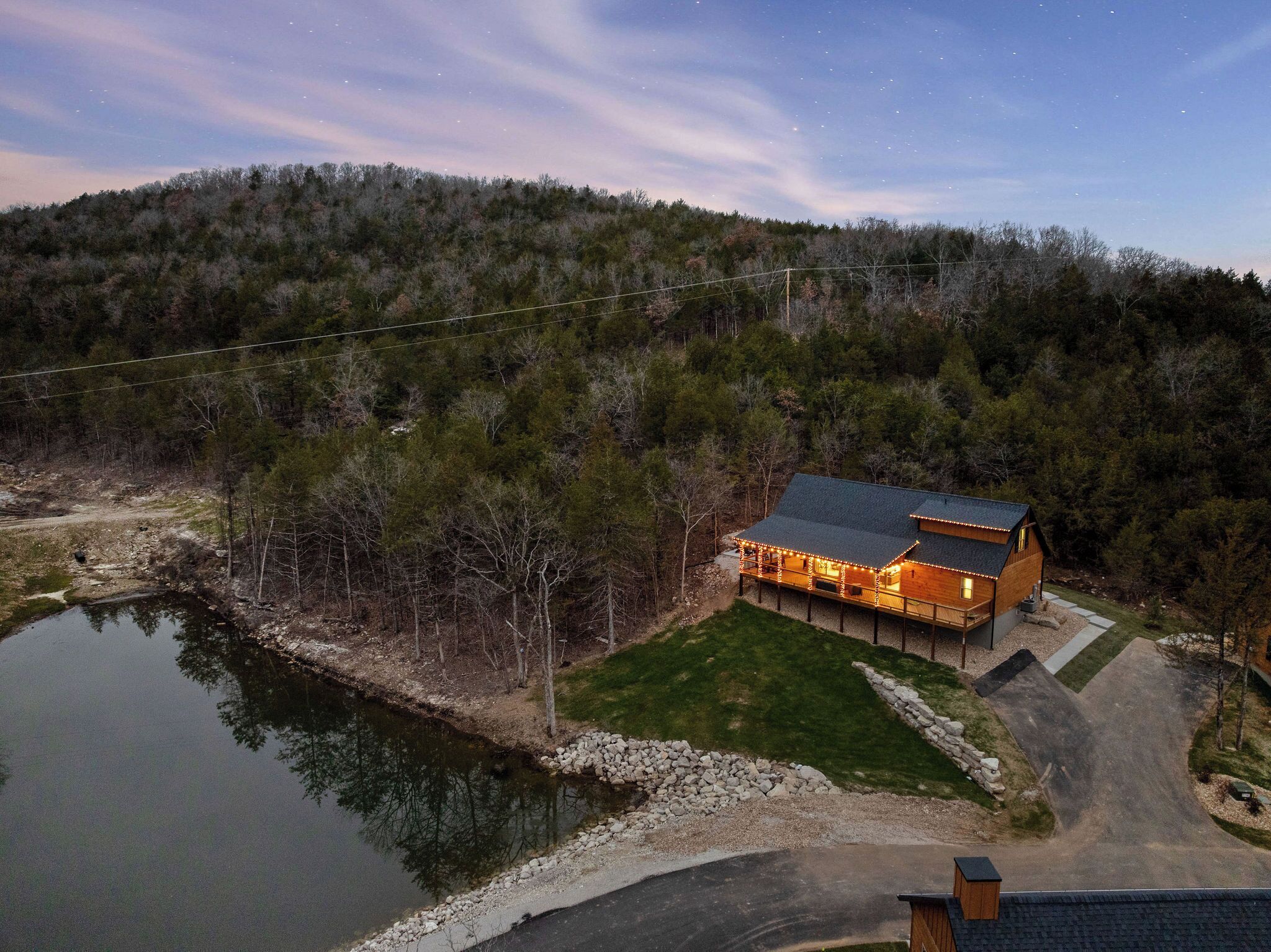 A cabin with a deck and lights, surrounded by trees and a body of water.
