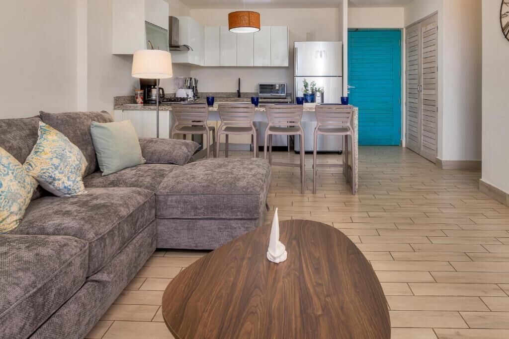 Uma sala de estar moderna com um sofá cinzento, uma mesa de centro de madeira e uma área de cozinha com armários brancos e uma porta azul.