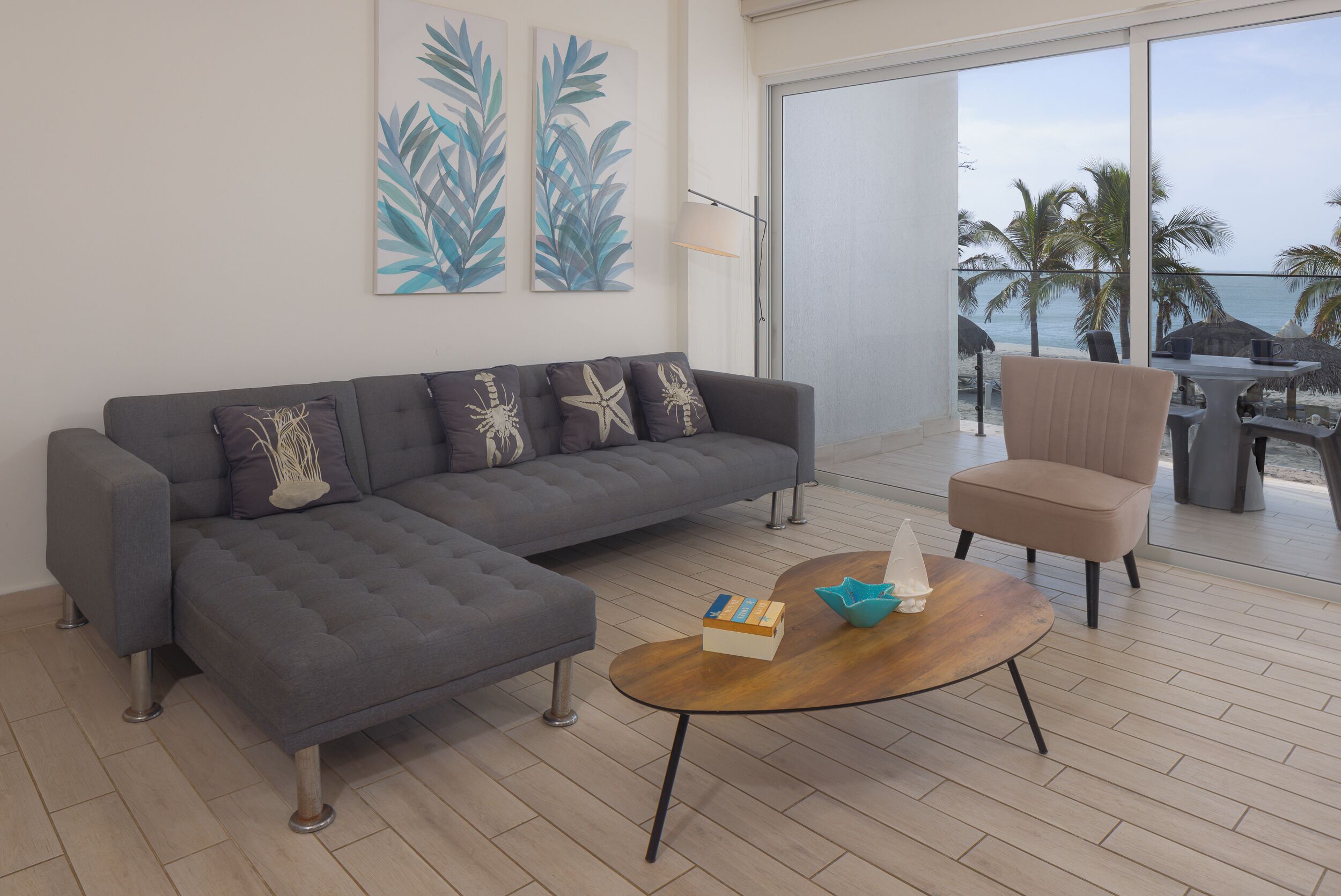 Een moderne woonkamer met een grijze modulaire bank, een houten salontafel en een beige fauteuil. Twee aan de muur bevestigde kunstwerken tonen tropische bladeren. De kamer heeft een grote schuifdeur die leidt naar een balkon met uitzicht op palmbomen en de oceaan.