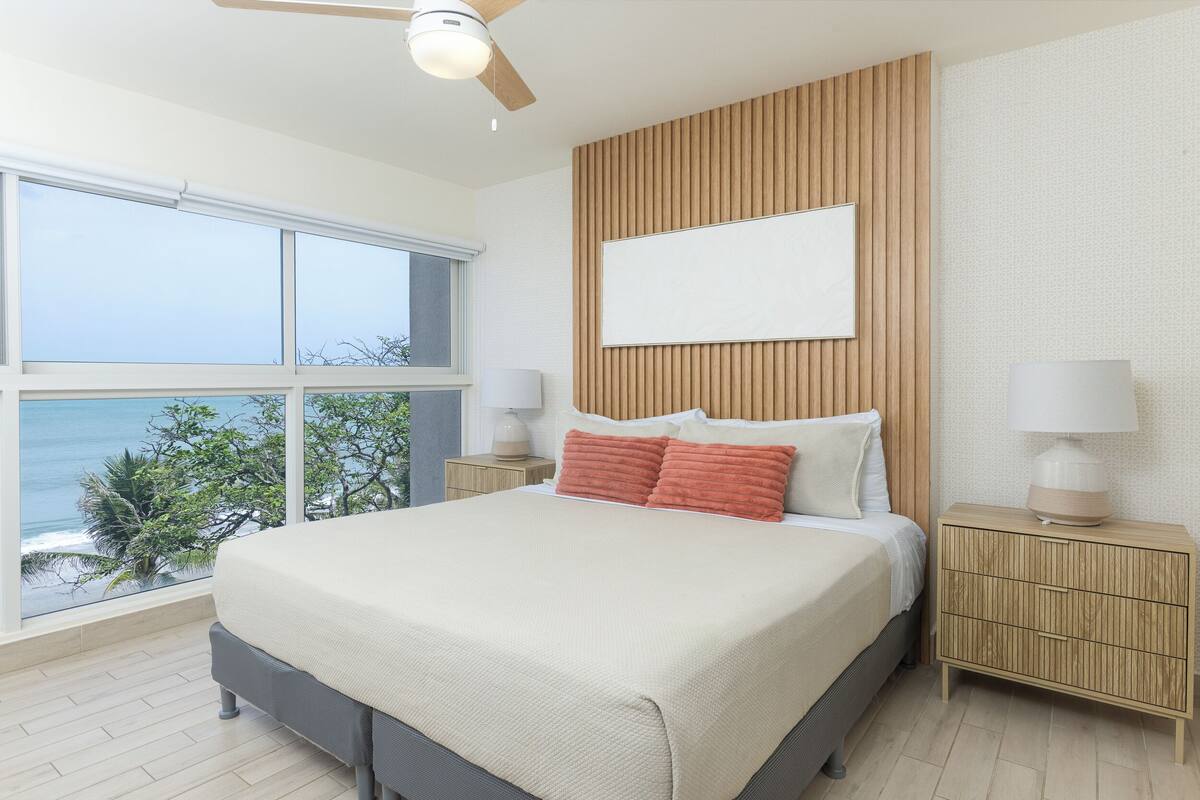 Een slaapkamer met een groot bed, een houten hoofdbord, twee nachtkastjes met lampen en uitzicht op de oceaan.