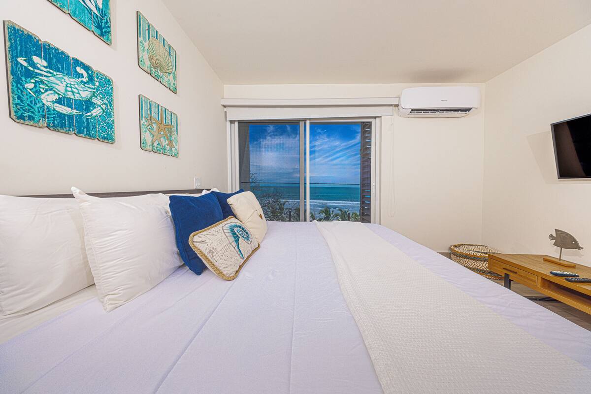 Een slaapkamer met een groot bed, uitzicht op de oceaan door een schuifdeur en een tv aan de muur.