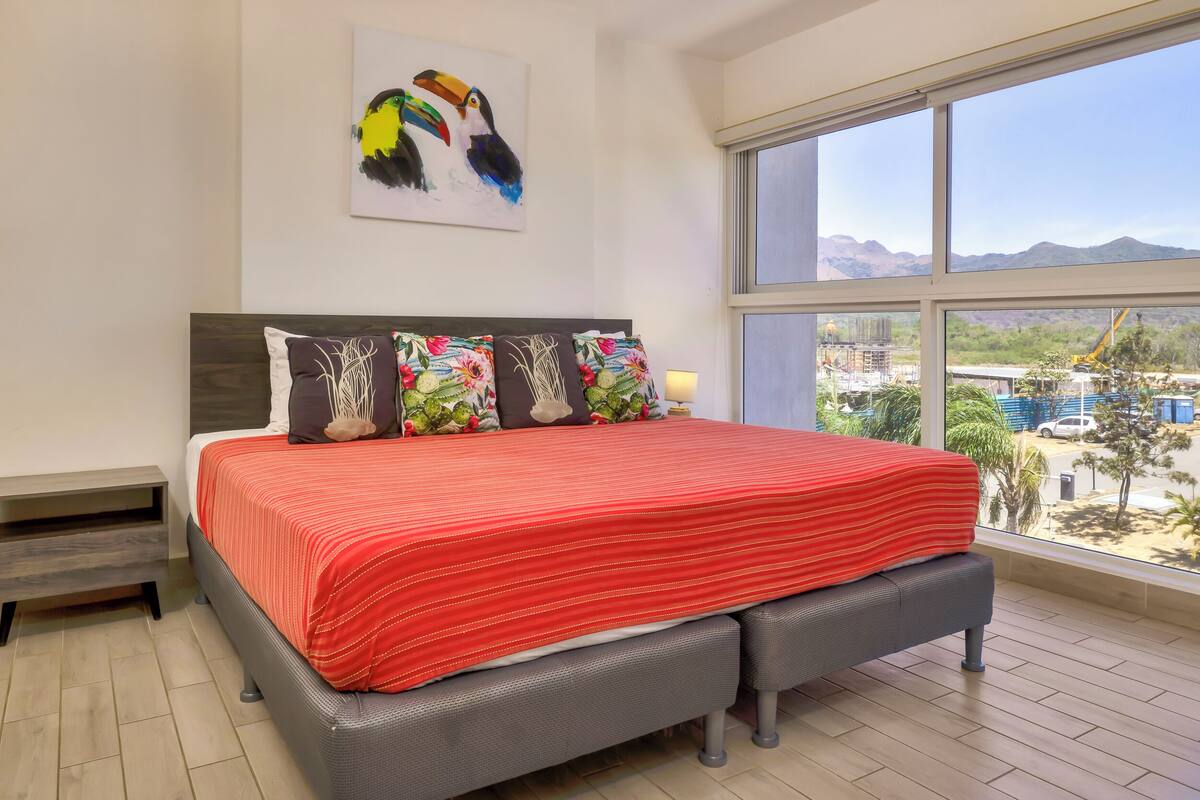 Slaapkamer met een groot bed, meerdere kussens, een nachtkastje en een raam met uitzicht op een bouwplaats.