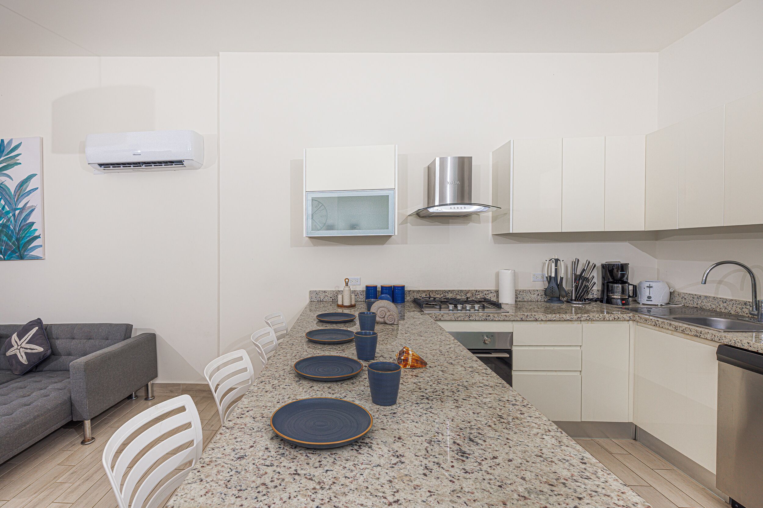 Une cuisine moderne avec des meubles blancs, un plan de travail en granit et un coin repas comprenant un canapé gris et des chaises blanches.