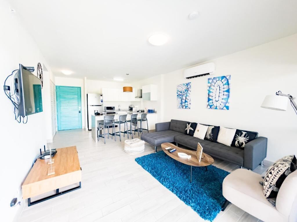 Una sala de estar moderna con un sofá gris, una mesa de centro de madera y una alfombra azul. Se observa una cocina al fondo con un comedor.