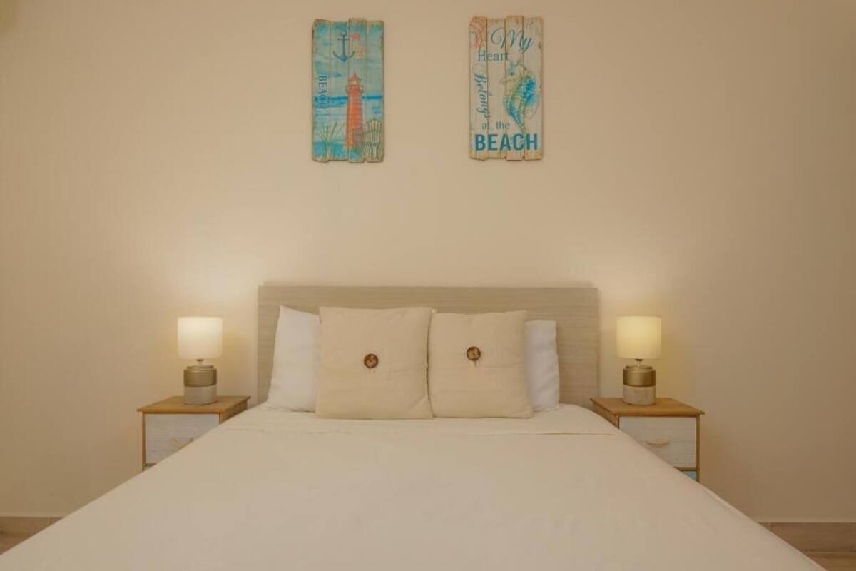 Een net opgemaakt bed met twee nachtkastjes, elk met een lamp, in een eenvoudige kamer met een lichtgekleurd hoofdeinde en effen muren.