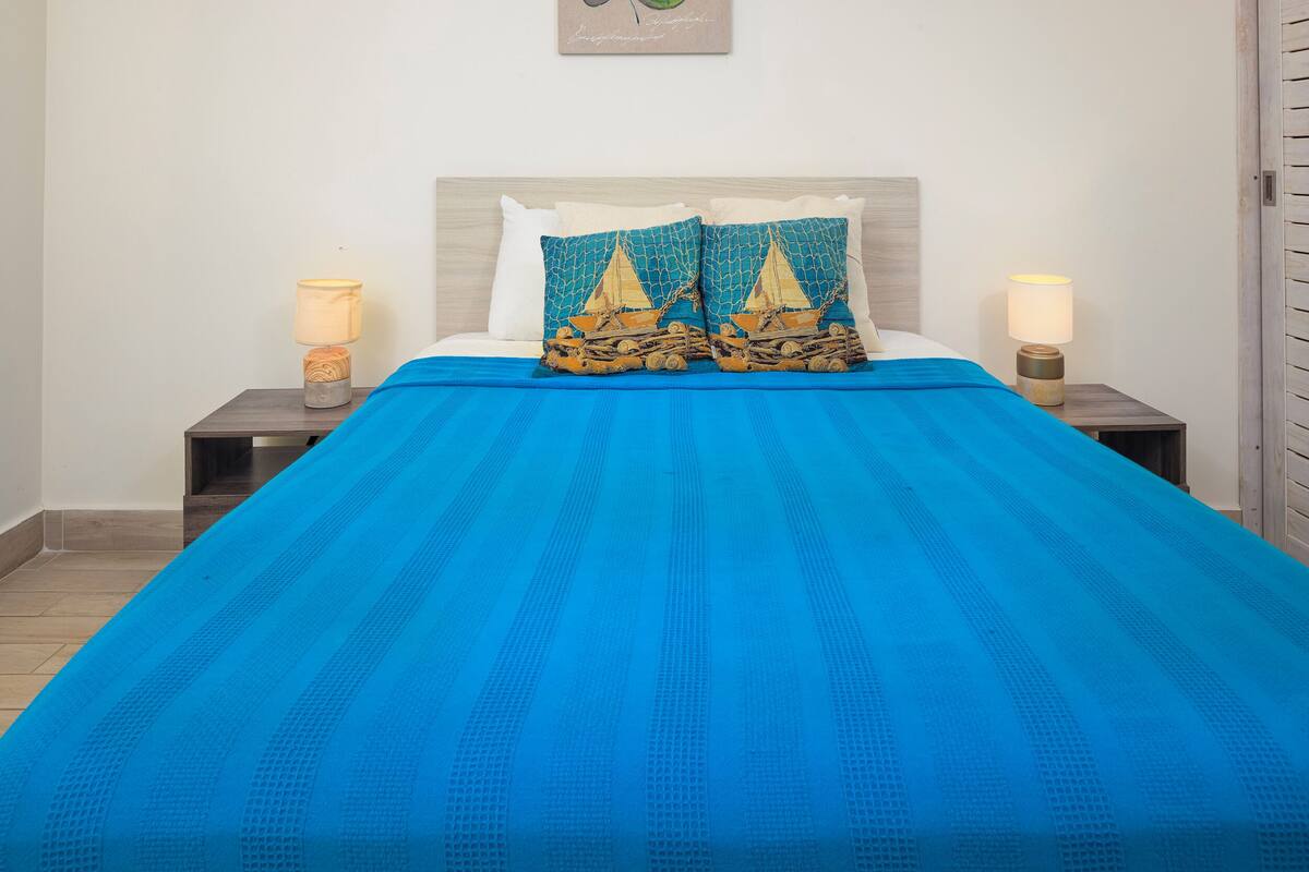 Een bed met een blauw dekbedovertrek, twee decoratieve kussens met een zeilbootmotief en twee bedlampen op houten nachtkastjes.