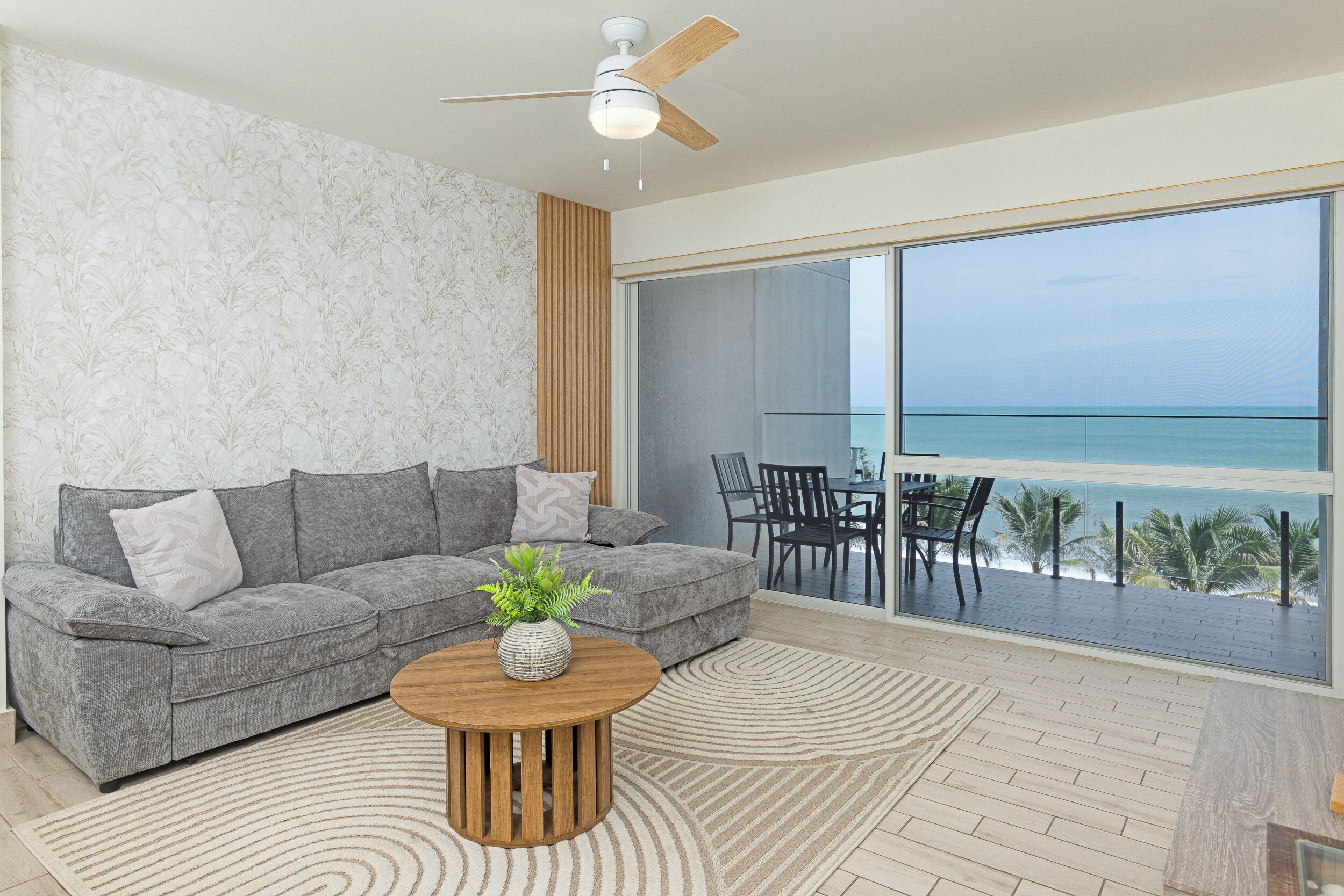 Un salón moderno con un sofá gris, una mesa de centro de madera y un gran ventanal con vistas a la playa.