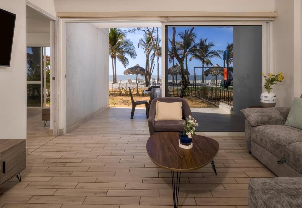Amplia sala de estar con piso de madera, un sofá, una mesa de centro y vista a la playa con palmeras y sombrillas.