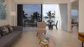 A modern living room with a sofa, a chair, and a coffee table. There is a large sliding glass door leading to a balcony with a view of the beach and palm trees.