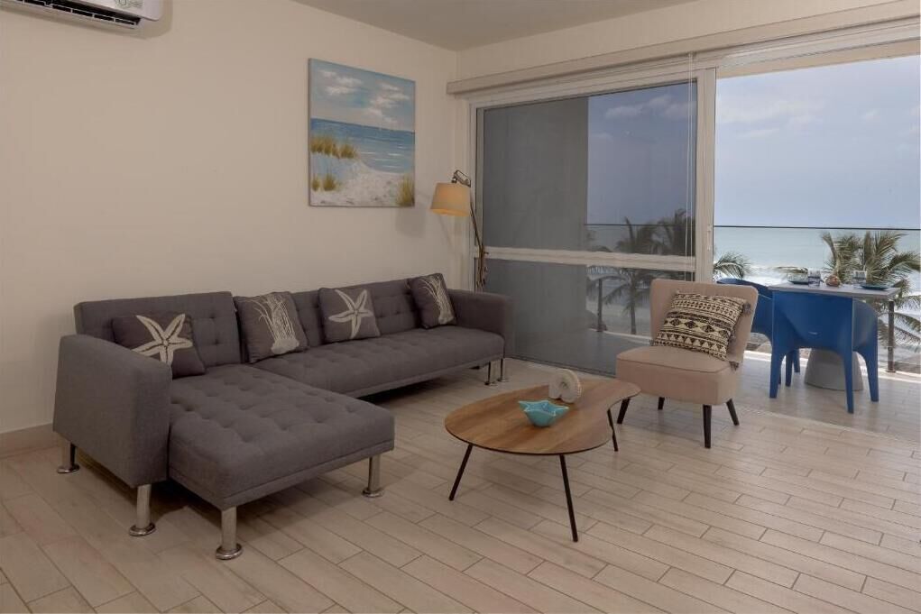 A living room with a grey sectional sofa, a round coffee table, and a beige armchair. There is a large window with a view of the ocean.