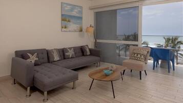 A living room with a grey sectional sofa, a round coffee table, and a beige armchair. There is a large window with a view of the ocean.