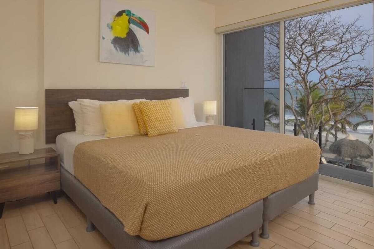 Een groot bed met een bruine deken, twee bedlampen, een houten nachtkastje en een schilderij van een vogel aan de muur.